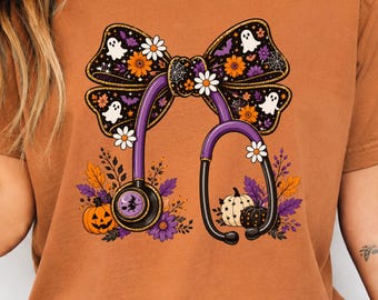 May include: A burnt orange t-shirt featuring a graphic of a purple stethoscope intertwined with a large black bow. The bow is decorated with white ghosts, orange flowers, and spiderwebs. Below, orange and white pumpkins sit among autumn leaves.
