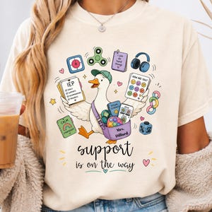 Puede incluir: Una camiseta de color crema con un gráfico de un pato que lleva una gorra verde y una bolsa morada. El pato está rodeado de varios objetos, como un fidget spinner, auriculares y una tarjeta con el texto "IEP" y "Mrs. Dillard". Debajo del pato, el texto dice "support is on the way".