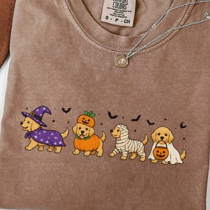 May include: A brown t-shirt featuring a graphic of four golden retriever puppies in Halloween costumes: a witch, a pumpkin, a mummy, and a ghost holding a jack-o'-lantern bucket. The shirt has a Comfort Colors label at the neck and a silver necklace.