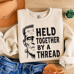 Op de afbeelding: Een crèmekleurige sweater met ronde hals voorzien van een zwarte afbeelding van de helft van het gezicht van Frankenstein's monster aan de linkerkant en de tekst "HELD TOGETHER BY A THREAD" in een druipend, vet lettertype rechts.