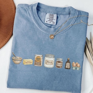 May include: A blue short-sleeved t-shirt with a graphic print of baking ingredients including a whisk, butter, brown sugar, milk, cocoa powder, spice, and eggs. The inner neck label reads Comfort Colors, 100% cotton, made in Honduras, size S.