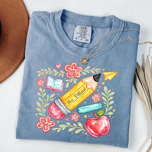 Puede incluir: Camiseta azul Comfort Colors con un diseño escolar. El diseño incluye un lápiz amarillo con "Mrs. Dillard", una manzana roja, flores, un libro y un avión de papel. Un collar dorado está sobre la camiseta.