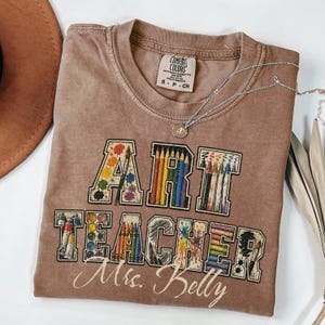 May include: A brown Comfort Colors t-shirt with the words "ART TEACHER Mrs. Betty" in a colorful design featuring paint splatters, paintbrushes, and pencils. A silver necklace with a pendant is draped across the shirt.