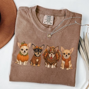 May include: A brown short-sleeve t-shirt featuring a graphic of four chihuahua dogs dressed in autumn-themed costumes, including a pumpkin, a sweater with an acorn, a turkey, and a scarf. The shirt has a Comfort Colors brand label at the neck.