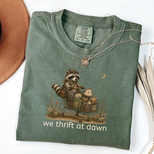 May include: A sage green t-shirt featuring a graphic of a raccoon wearing a hat and scarf, pushing a shopping cart filled with various items. The text "we thrift at dawn" is printed below the graphic. The shirt has a Comfort Colors brand label.