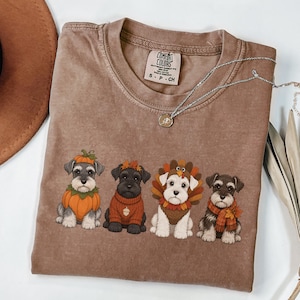 Puede incluir: Una camiseta marrón de manga corta con un gráfico de cuatro perros Schnauzer vestidos con disfraces de temática otoñal, incluyendo una calabaza, un suéter con una bellota, un pavo y una bufanda de cuadros. Etiqueta de marca Comfort Colors.