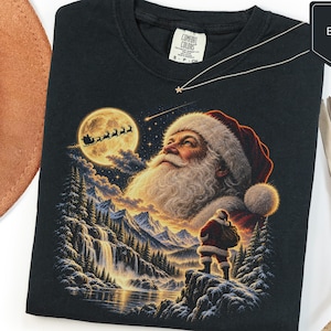 Puede incluir: Una camiseta negra con un estampado gráfico de Santa Claus mirando hacia una luna llena con la silueta de un trineo. Debajo, una figura pequeña de Santa Claus se encuentra en un acantilado nevado sobre un paisaje montañoso con cascada.