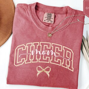 May include: A crimson short-sleeve t-shirt with a crew neckline and a Comfort Colors label. The front features the text CHEER in a large, cream-coloured collegiate font, with the word mom in a cursive script and a small bow graphic below.