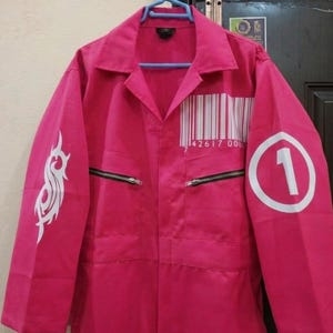 Puede incluir: Mono rosa brillante con cuello y cierre de cremallera. La manga izquierda presenta un diseño tribal blanco, y la manga derecha muestra un número uno blanco dentro de un círculo. Un código de barras y números están impresos en el pecho.