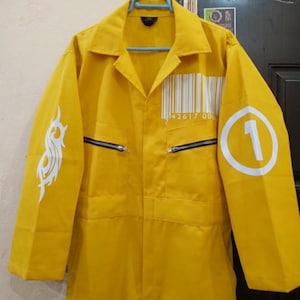 Peut inclure: Combinaison jaune vif avec un col et des manches longues. Comprend un code-barres blanc et le chiffre "1" dans un cercle blanc sur les manches. Comprend des motifs tribaux blancs sur la manche gauche et des fermetures éclair noires.