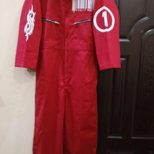 Peut inclure: Combinaison rouge avec col, fermeture éclair et manches longues. Comprend un motif blanc sur la manche gauche et un numéro 1 blanc dans un cercle sur la manche droite. Comprend un code-barres et des chiffres sur la poitrine.