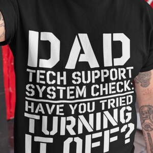 May include: Black t-shirt with white text: "DAD TECH SUPPORT SYSTEM CHECK: HAVE YOU TRIED TURNING IT OFF?" The text is in a bold, sans-serif font. The shirt is a crew neck and appears to be made of cotton.