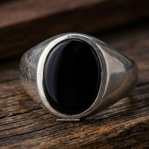 May include: A silver signet ring featuring a smooth, polished oval black stone set in the center. The metal band has a brushed finish and the text jewelrybycrown is printed in small white letters at the base of the ring setting.