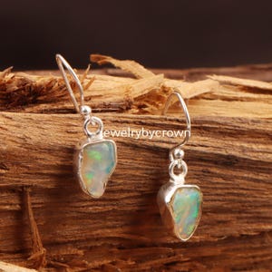 Könnte beinhalten: Silberne Ohrringe mit tropfenförmigen Opalsteinen. Die Opale zeigen eine Mischung aus Farben, darunter Grün, Weiß und Gelb. Die Ohrringe sind vor einem rustikalen Holzgrund platziert.