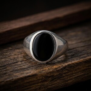 Peut inclure: Une bague en argent avec une grosse pierre noire ovale sertie dans un anneau en métal brossé. La bague est présentée sur une surface en bois, mettant en valeur son design poli. Les bijoux sont de jewelrybycrown.