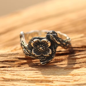 Könnte beinhalten: Ein silberner Ring mit floralem Design. Der Ring zeigt eine detaillierte Blume mit strukturierter Oberfläche, die auf einem Band angebracht ist, das einen Ast nachahmt. Der Ring wird auf einer Holzoberfläche mit dem Text "jewelrybycrown" angezeigt.