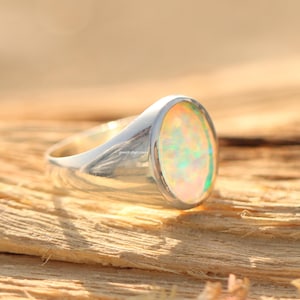 Può includere: Un anello d'argento con una pietra preziosa di opale ovale. L'opale mostra una miscela di colori, tra cui giallo, verde e rosa. L'anello è posto su una superficie di legno, con il logo jewelrybycrown visibile.