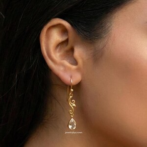 May include: Gold-toned drop earring with a teardrop-shaped gemstone. The earring features a decorative, swirling design and a hook closure. The gemstone is clear and faceted. The text "jewelrybycrown" is visible.
