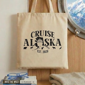 May include: A beige tote bag with the words "CRUISE ALASKA" in black, along with an image of a bear and other wildlife. The text "EST. 2026" is below. The bag hangs on a hook near a round window with a mountain view.