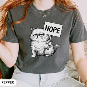 Może przedstawiać: Ciemnoszara koszulka z wściekłym kotem z kresk&oacute;wki trzymającym znak z napisem "NOPE". Kot ma surowy wyraz twarzy i jest narysowany w szczeg&oacute;łowym, szkicowym stylu. Koszulka jest w stylu casual.