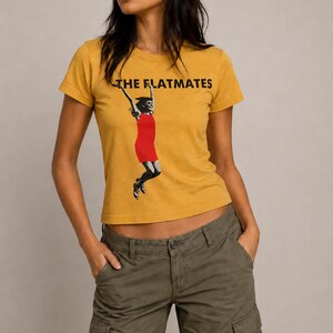 May include: A mustard yellow short-sleeve t-shirt featuring the black text THE FLATMATES and a graphic of a person jumping while wearing a red dress. The shirt has a crew neckline and a fitted silhouette, paired with olive green cargo pants.