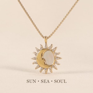 May include: Gold-toned necklace featuring a sun and moon pendant. The pendant has a crescent moon with a face and a sunburst design, adorned with small crystals. The necklace includes the text "SUN • SEA • SOUL".