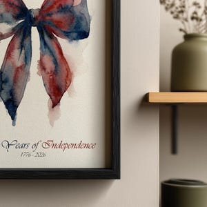 May include: Watercolor print of a bow in shades of red and blue, framed in black. The artwork is titled "Years of Independence" with the dates 1776-2026 in a decorative font. The bow is painted with watercolor techniques.