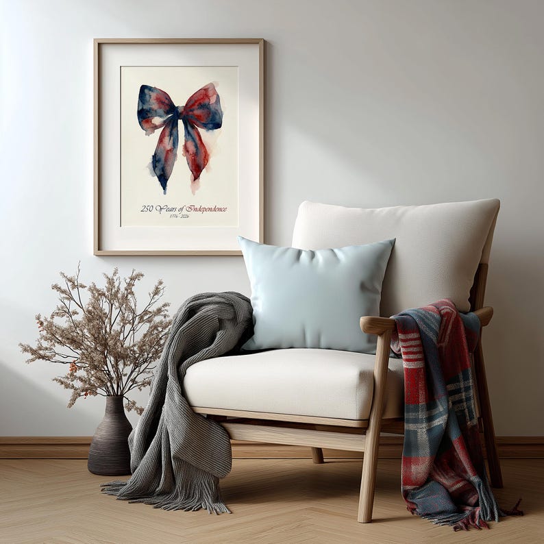 May include: A framed watercolor print of a bow in red and blue hues, with the text "250 Years of Independence 1776-2026." The print hangs above a cream-colored armchair with a light blue pillow and a plaid throw.