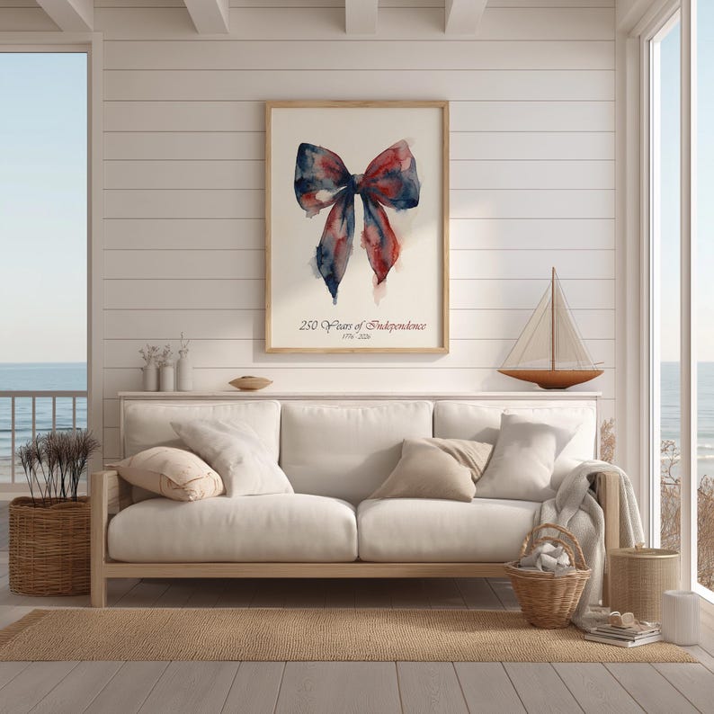 May include: A framed watercolor print featuring a bow in shades of red and blue. The print is displayed above a white sofa in a bright, airy room. The text "250 Years of Independence 1776-2026" is below the bow.