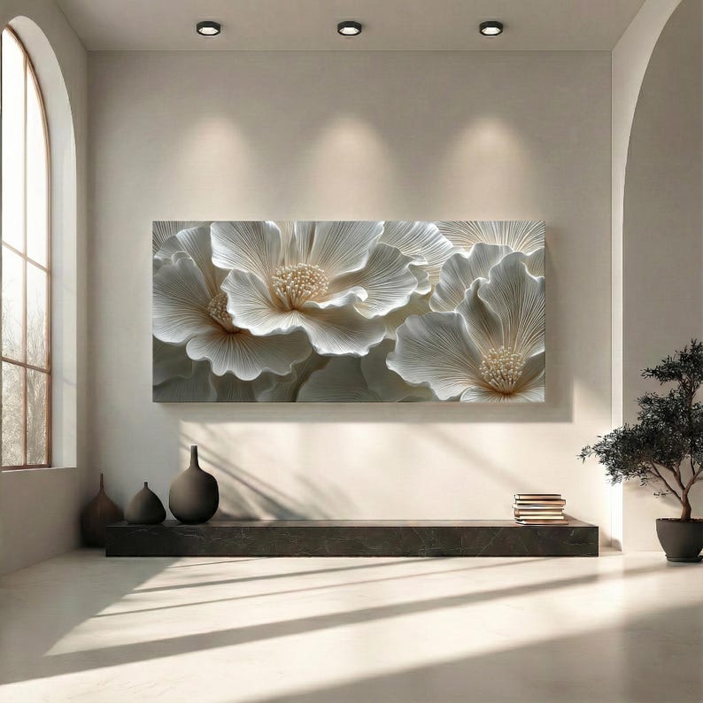May include: A large, rectangular wall art piece featuring a relief of three white flowers with textured petals and golden centers. The artwork is mounted on a neutral-toned wall, creating a serene and elegant atmosphere. The room is minimalist, with a dark shelf and decorative vases.