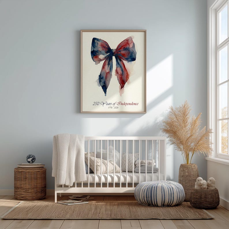 May include: Watercolor painting of a bow in shades of red and blue, framed in a light wood. The text "250 Years of Independence (1776-2026)" is below the bow. The artwork hangs above a white crib in a light blue room.