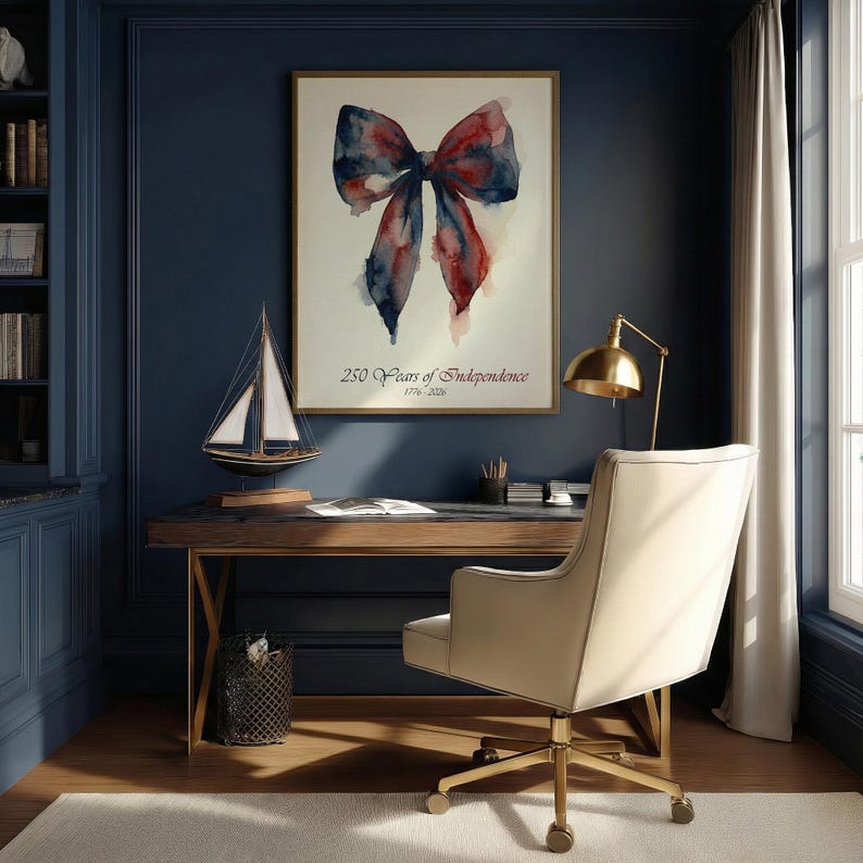 May include: A framed watercolor painting of a red and blue bow hangs above a desk. The text "250 Years of Independence 1776-2026" is below the bow. A model sailboat and a gold desk lamp are on the desk. A cream-colored chair sits in front of the desk.