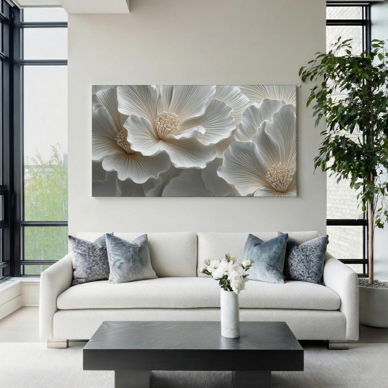 May include: A large, rectangular wall art piece featuring a close-up of white, textured flowers with beige centers. The artwork hangs above a white sofa with blue pillows. A black coffee table and a vase of white flowers complete the scene.