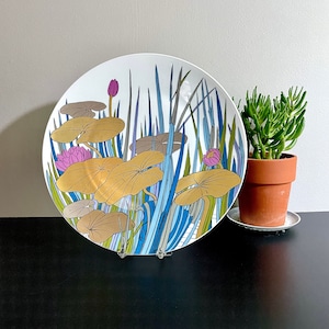 Puede incluir: Un plato de cer&aacute;mica blanco con un dise&ntilde;o azul, verde y dorado de nen&uacute;fares y juncos estilizados. El plato est&aacute; sobre una superficie negra.