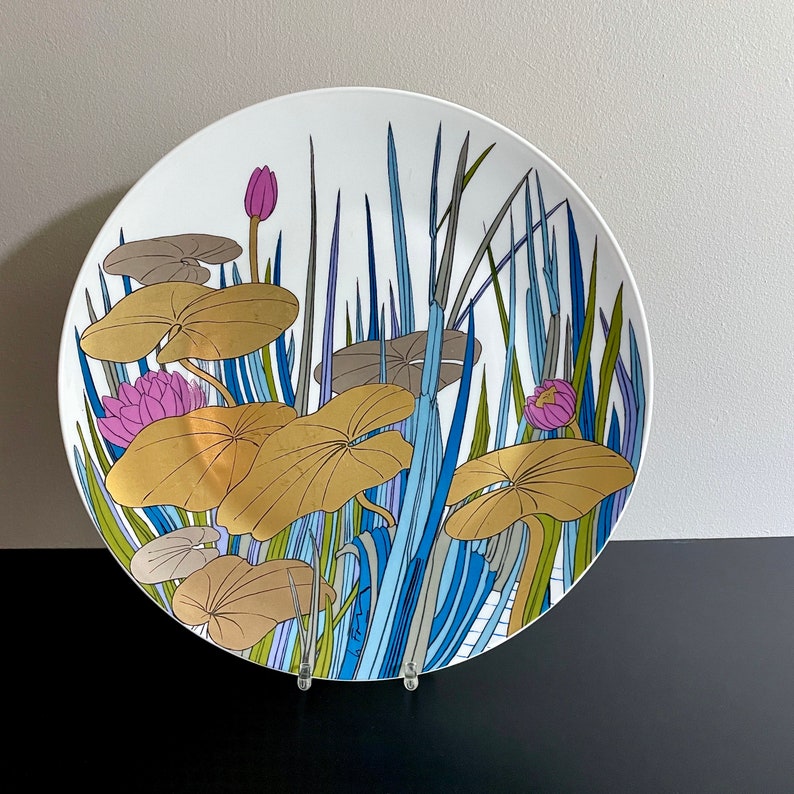 Puede incluir: Un plato de cer&aacute;mica blanco con un dise&ntilde;o colorido de nen&uacute;fares y hojas azules, verdes y dorados. El dise&ntilde;o es estilizado y abstracto.