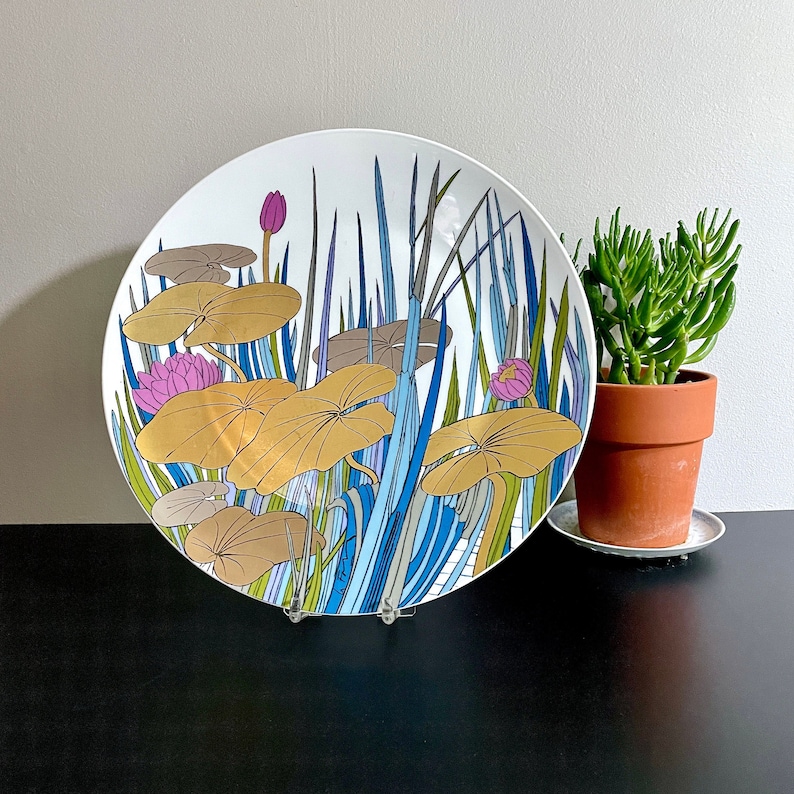 Puede incluir: Un plato de cer&aacute;mica blanco con un dise&ntilde;o azul, verde y dorado de nen&uacute;fares y juncos estilizados. El plato est&aacute; sobre una superficie negra.