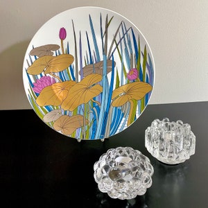 Puede incluir: Un plato de cer&aacute;mica blanco con un dise&ntilde;o floral azul y verde con nen&uacute;fares y hojas estilizados. El plato est&aacute; sobre una superficie negra con dos candelabros de vidrio transparente.