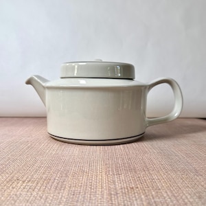 May include: A light gray ceramic teapot with a flat lid and a curved handle. The teapot features a simple, low-profile cylindrical body with a short spout and a thin, dark brown horizontal line accenting the base of the vessel.