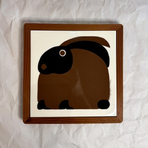 May include: Square ceramic tile with a brown and black rabbit illustration, set in a wooden frame. The rabbit has a black head and ears, a white eye, and a brown body. The tile is on a white background.