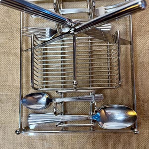 May include: A chrome-plated flatware organizer with a rectangular frame. The organizer holds forks, knives, and spoons. The utensils have a ribbed handle design. The organizer is designed to sit on a table or countertop.