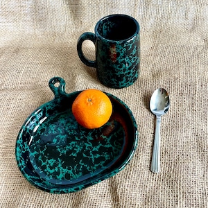May include: A green and black ceramic plate with a handle, a green and black ceramic mug, and a silver spoon. An orange sits on the plate.