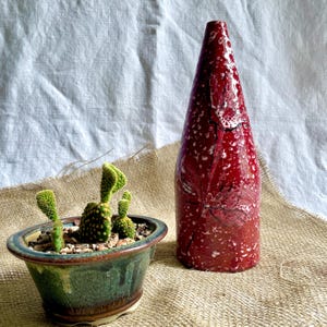 May include: A red ceramic vase with a white speckled glaze and a raised floral design. The vase is tall and narrow, with a rounded base and a pointed top. It is sitting on a burlap surface next to a small green pot with a cactus plant.