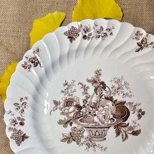 May include: White ceramic serving platter with a brown floral and fruit basket design. The rim has a scalloped edge with a repeating floral pattern. The platter is a vintage style and is suitable for serving food.