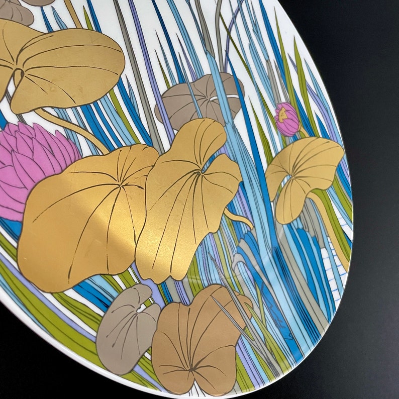 Puede incluir: Un plato de cer&aacute;mica blanco con un dise&ntilde;o floral dorado y azul. El dise&ntilde;o presenta hojas y flores estilizadas en tonos dorados, azules, verdes y rosas.