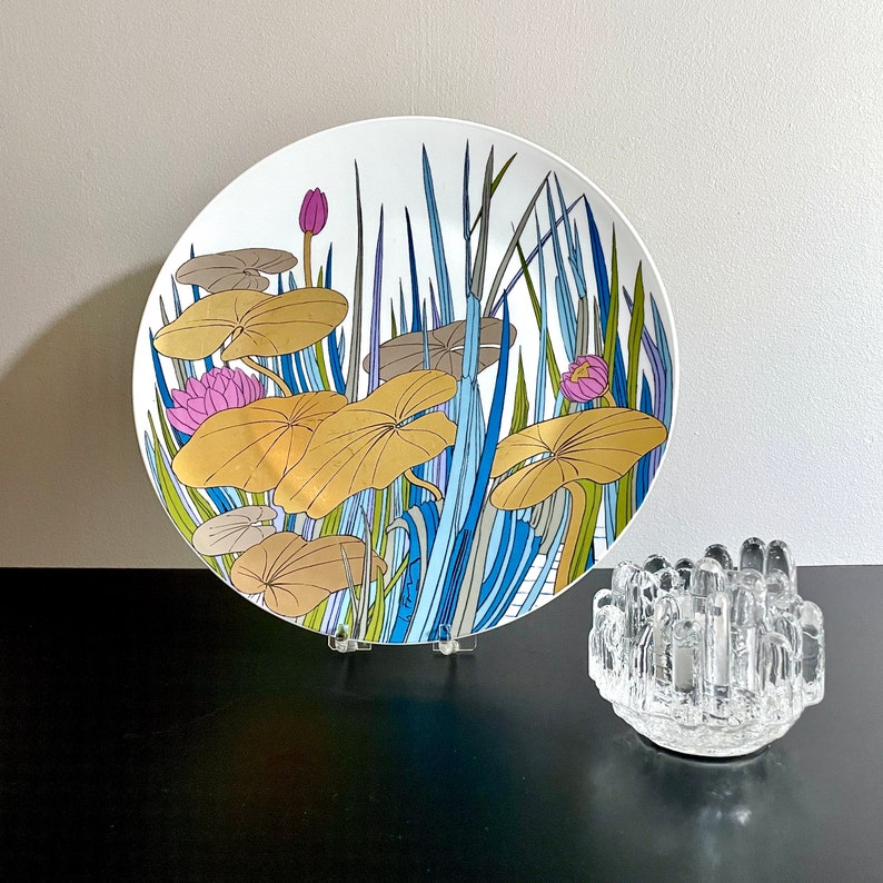 Puede incluir: Un plato de cer&aacute;mica blanco con un dise&ntilde;o floral azul, verde y dorado. El dise&ntilde;o presenta nen&uacute;fares y juncos estilizados.