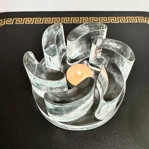May include: A clear glass candle holder with six curved sections arranged in a circular pattern. A lit tea light candle sits in the center. The candle holder is on a black surface.