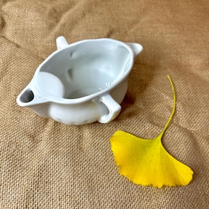 May include: A white ceramic gravy boat with a spout and two handles. A bright yellow ginkgo leaf rests beside the boat on a tan burlap surface. The gravy boat has a smooth, glossy finish and a classic design.