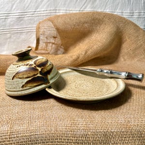 May include: A ceramic butter dish with a speckled cream glaze and a domed lid featuring dark brown leaf-like patterns. The set includes a matching circular base plate and a silver-toned metal butter knife with a decorative bamboo-style handle.