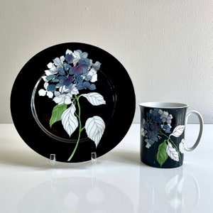 May include: A black ceramic plate and mug set featuring a floral hydrangea design. The plate has a wide rim and the mug has a white handle. The hydrangea design includes blue, white, and green colors.