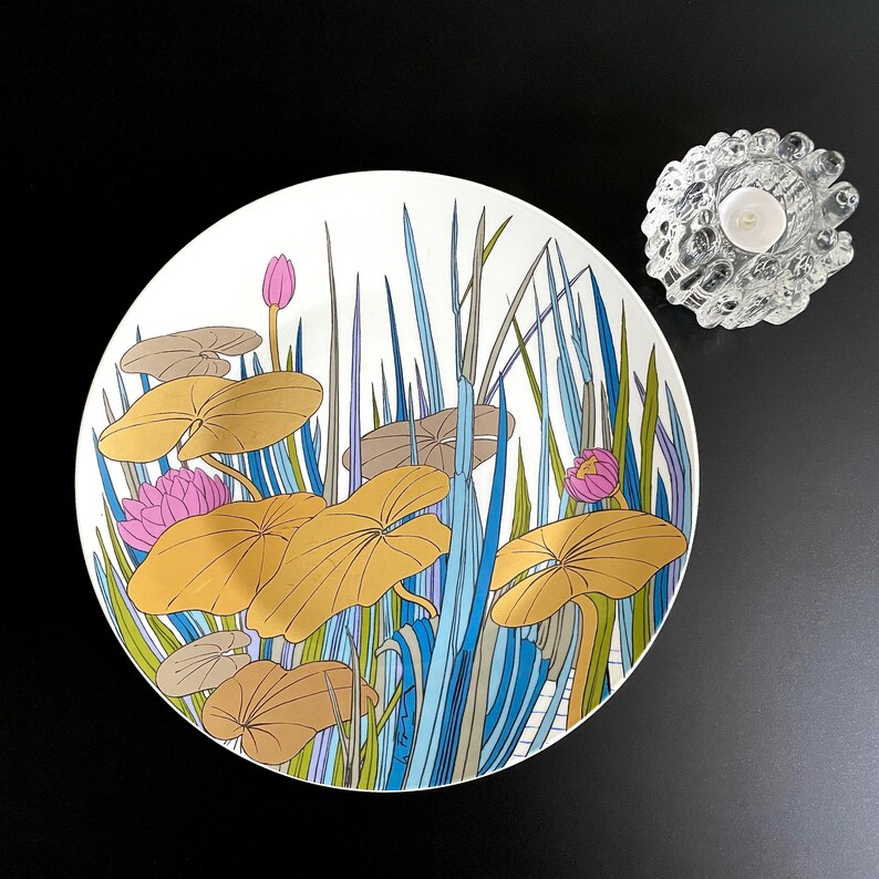 Puede incluir: Un plato de cer&aacute;mica blanco con un dise&ntilde;o floral azul, verde y rosa. El dise&ntilde;o presenta nen&uacute;fares y hojas estilizados.